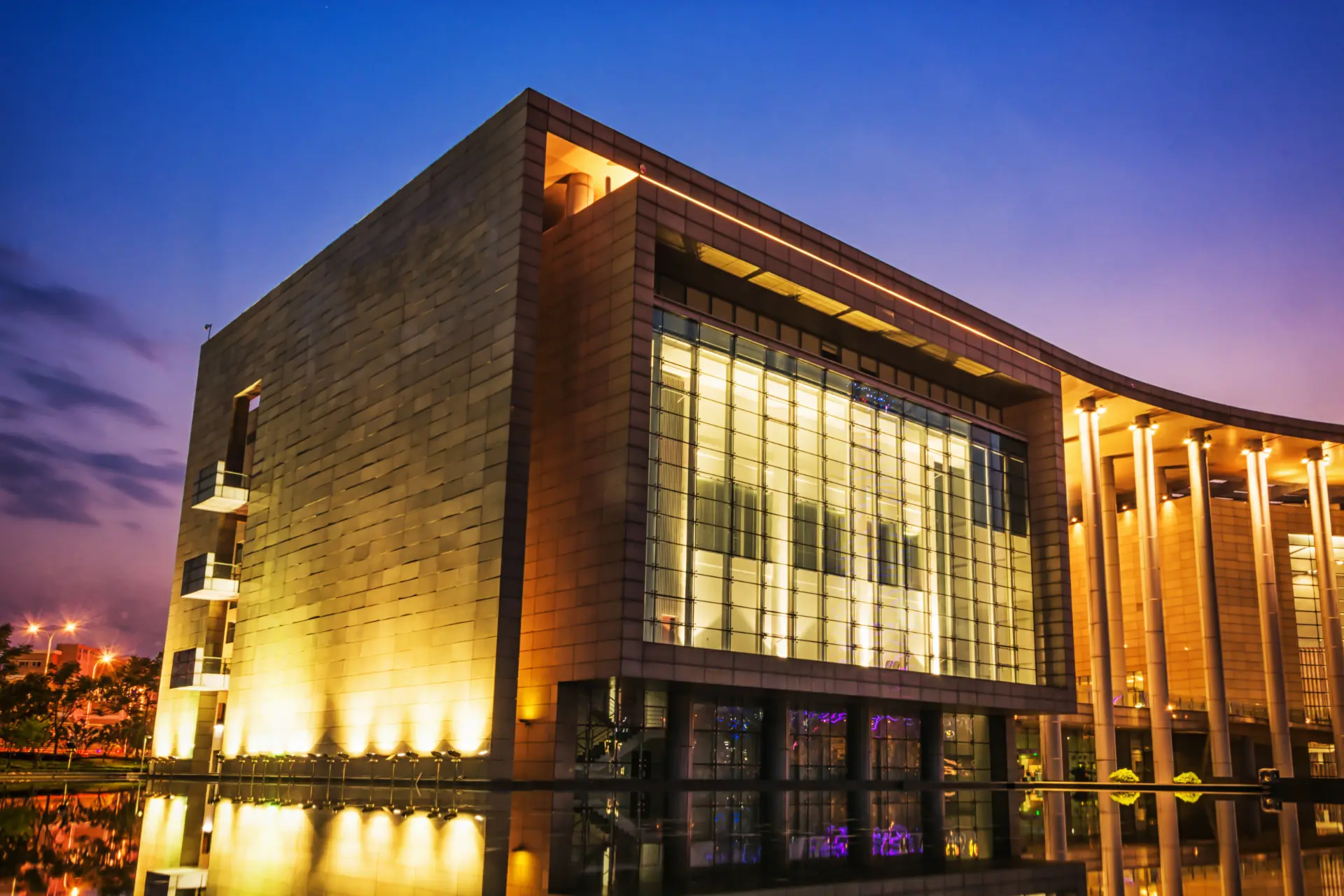 At dusk, a modern glass-and-stone building glows with architectural lighting, reflected in the water below.