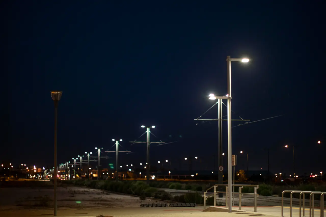 At night, modern streetlights illuminate a paved walkway, with rows of lamps receding toward distant roads.