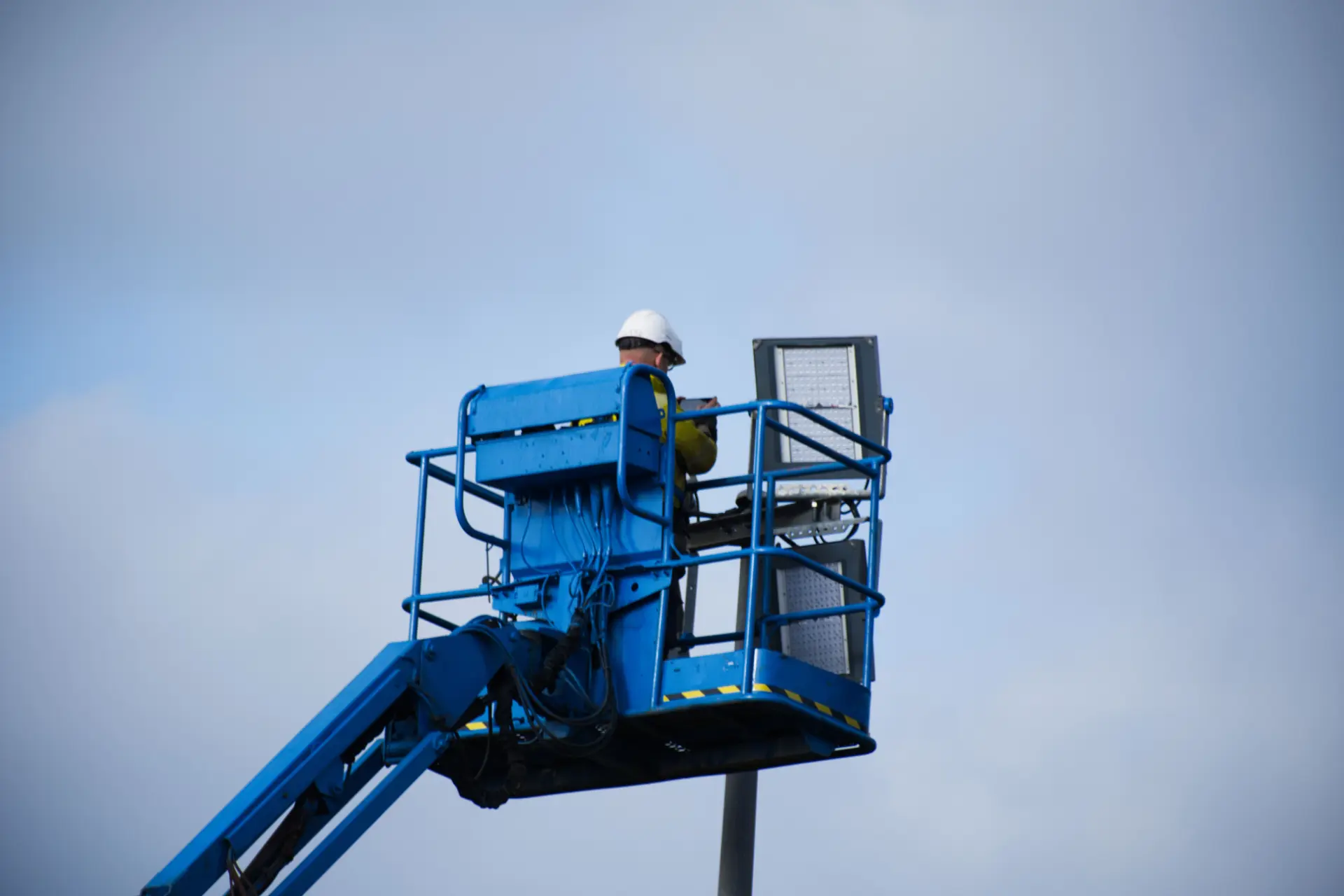 Ein Monteur wartet eine Straßenleuchte auf einem Mast und arbeitet dafür von einer blauen Hebebühne aus hoch oben.